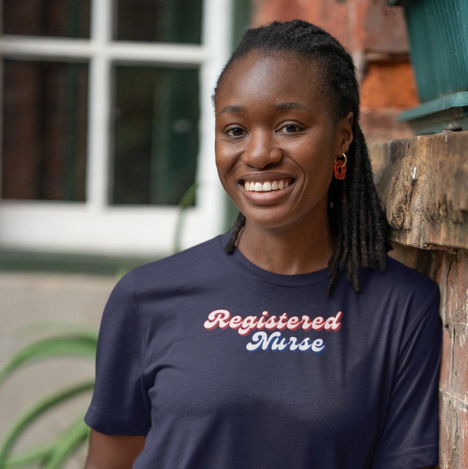 Woman wearing a navy blue t-shirt with 'Registered Nurse' text, standing against a brick wall.