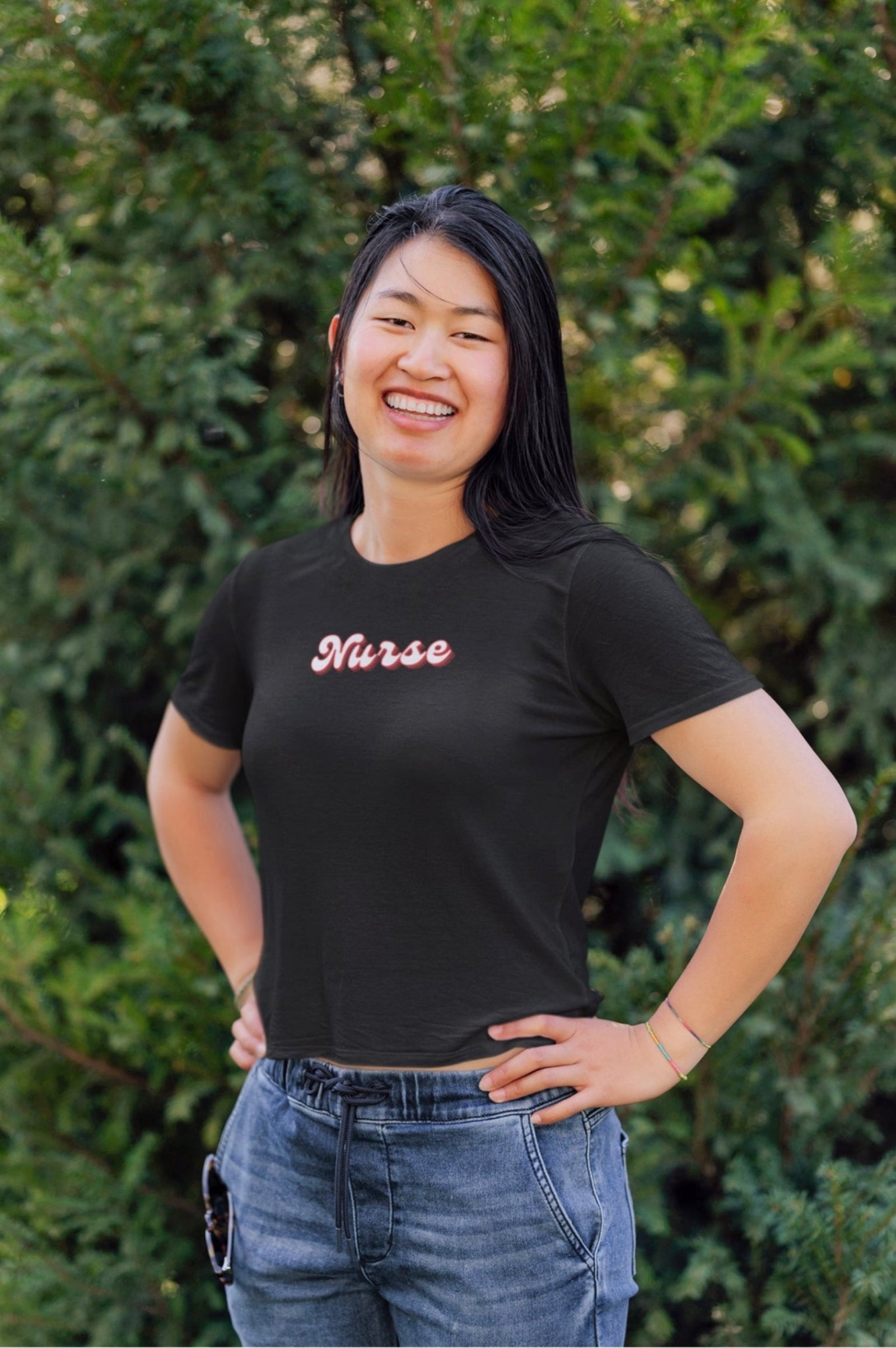 Woman wearing a black t-shirt with 'Nurse' printed on it, standing outdoors with greenery in the background.